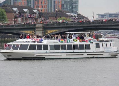 The Sarpedon, a white river boat with flags along its upper rail, passing a Thames bridge by day