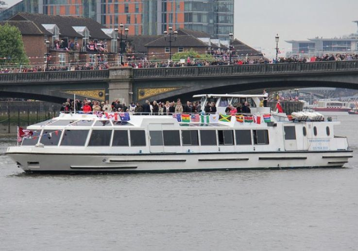 The Sarpedon, a white river boat with flags along its upper rail, passing a Thames bridge by day