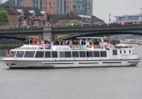 The Sarpedon, a white river boat with flags along its upper rail, passing a Thames bridge by day